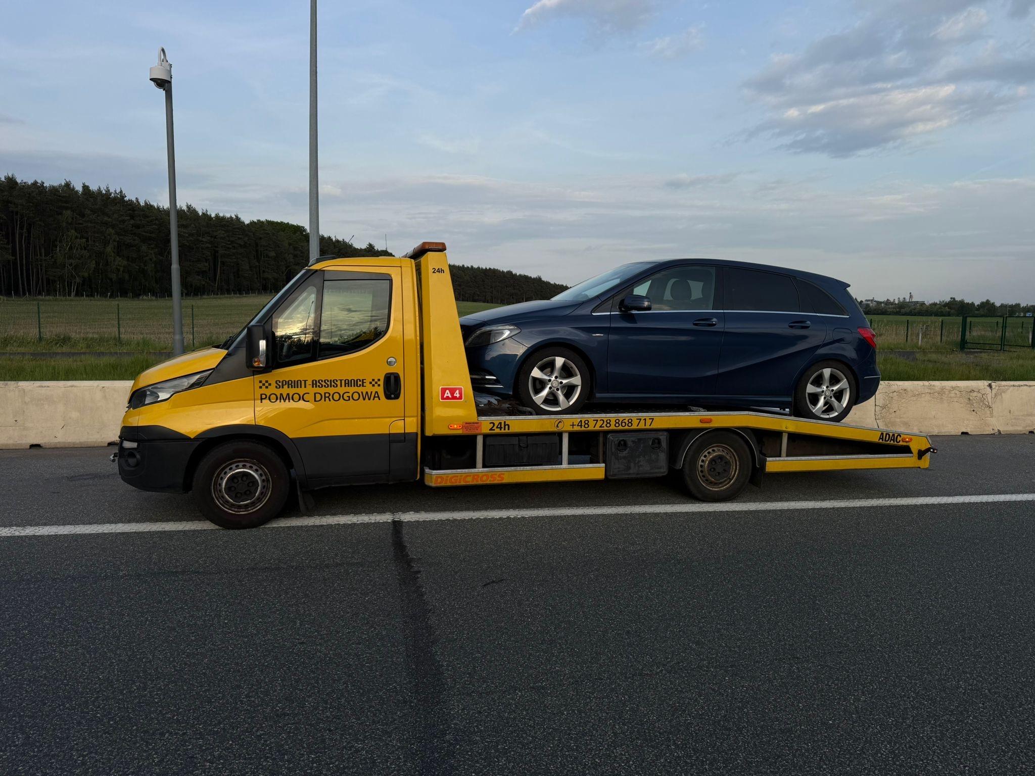 Laweta z granatowym Mercedesem na platformie, zatrzymana na pasie awaryjnym autostrady